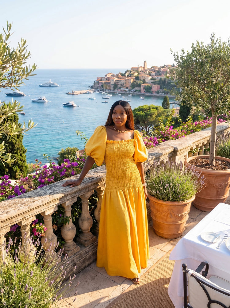 Woman in a yellow dress standing on a balcony overlooking a scenic coastal view with boats and buildings.
